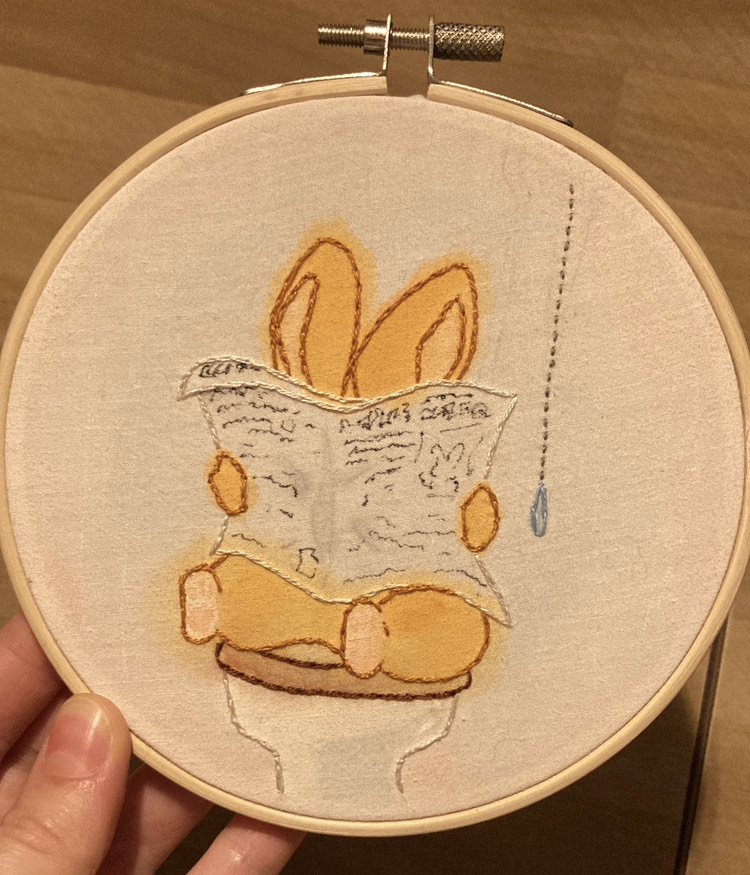 hoop embroidery with light pink watercolour background; a small ginger bunny is sitting on an old-timey toilet, mostly covered up by a newspaper his little arms are nearly too short to hold properly.